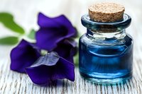 Small glass bottle with blue liquid and cork stopper next to vibrant purple flower petals on rustic white wooden surface, macro photography for spa and wellness.