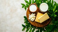 Fresh dairy products on wooden board with green leaves frame, milk glasses, butter block and cheese cubes on light marble background. Food photography.