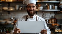Smiling bearded male chef in white uniform and hat holding blank white board in restaurant kitchen. Professional culinary background for menu or cooking advertisement.