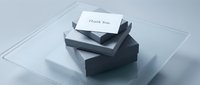 Elegant thank you card resting on stacked gray gift boxes on a glass tray, minimalist style for corporate gifting and appreciation concepts.