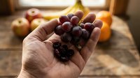Red grapes held in open palm with fresh fruit assortment blurred in background, suitable for healthy eating, nutrition, and organic produce marketing.