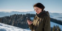 Young woman in green winter jacket using smartphone on snowy mountain peak with forest and misty mountain ranges in background.