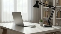 Modern minimalist home office workspace with laptop computer and adjustable desk lamp on white table near window with natural light for productivity.