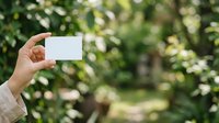 Hand holding blank white card outdoors with green foliage background, suitable for business card mockups, branding presentations, and marketing material displays.