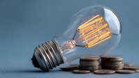 Vintage Edison bulb with glowing filament next to stack of coins on blue background representing energy costs and electricity savings concept.