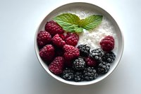 Fresh ripe raspberries and blackberries in white ceramic bowl with creamy yogurt and mint leaf garnish on light background, healthy breakfast concept.