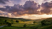 Dramatic sunset over rolling green hills and valleys with golden light breaking through storm clouds, suitable for landscape and nature backgrounds.