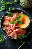 Prosciutto and cantaloupe melon appetizer with fresh basil leaves served on dark ceramic plate, gourmet Italian antipasto for summer dining and entertaining.