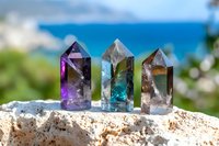 Three colorful crystal points in purple, blue and clear quartz arranged on natural stone with ocean backdrop for healing and meditation practices.