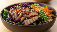 Grilled teriyaki chicken breast sliced over fresh salad with purple cabbage, shredded carrots, spinach and sesame seeds in dark ceramic bowl.