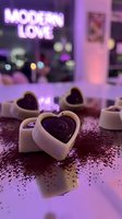 Heart-shaped chocolates in white ceramic holders with dark ganache centers on glittery surface, neon modern love sign glowing in purple backdrop, romantic gift concept.