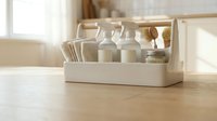 White wooden caddy with glass milk bottles and kitchen utensils on light wood table in bright modern kitchen interior for home organization and storage.