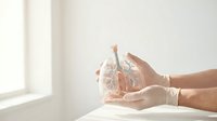 Hands in medical gloves holding glass anatomical heart model in bright clinical setting for healthcare education and medical training purposes.