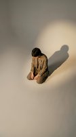 Young man with dark hair, South Asian ethnicity, wearing tan blazer and trousers, kneeling on white floor with dramatic shadow cast on wall behind, contemplative pose for mental health, stress, anxiety, depression, or introspection themes.