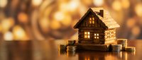Wooden house model surrounded by golden coins on warm bokeh background representing real estate investment and property savings concept.