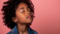 Young African American woman with natural curly hair and closed eyes expressing serenity against pink background, wearing denim jacket. Peaceful mindful portrait.