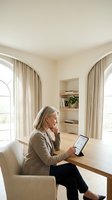 Mature Caucasian businesswoman working on tablet computer at home office desk in elegant beige interior with natural light from arched windows.