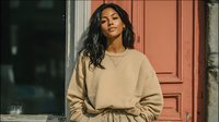 Young Hispanic woman in beige oversized sweater standing by red wooden door, casual autumn fashion portrait for lifestyle and clothing concepts.