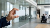 Businessman holding blank white business card in modern office lobby with glass walls and blurred colleagues in background, ready for contact information or branding.