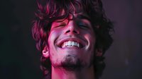 Young Caucasian man with curly dark hair and facial stubble laughing genuinely with eyes closed, illuminated by vibrant pink and purple neon lighting in dark atmosphere.