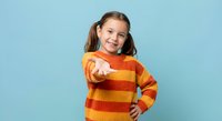 Happy young girl in striped orange sweater pointing finger at camera with confident smile on blue background for advertising and promotional content.
