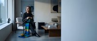Woman in black clothing sitting on floor with blue and yellow striped fabric in modern minimalist bedroom interior with natural light from window.