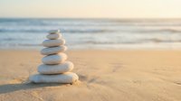 Zen stone stack balanced on sandy beach with soft ocean waves in background creating peaceful meditation and mindfulness atmosphere for wellness concepts.