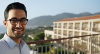 Young man with dark hair and glasses wearing light blue shirt smiling at camera on residential balcony with mountain views and apartment building in background.