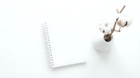 Minimalist workspace with blank spiral notebook and cotton branches in white vase on clean white background for creative projects and planning.