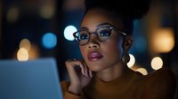 Professional African American businesswoman wearing glasses working late at night on laptop with city lights bokeh background for corporate presentations.