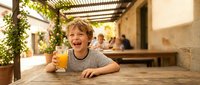 Happy young boy enjoying fresh orange juice at outdoor restaurant terrace during sunny day. Child smiling while drinking healthy beverage on vacation.