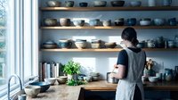 Young Asian woman potter working in ceramic studio with handmade bowls displayed on wooden shelves. Artisan crafting pottery in modern workshop space.