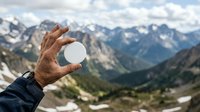 Weathered male hand holding white sphere against alpine mountain valley landscape with forested slopes and snow-capped peaks, outdoor adventure and nature concept for travel and exploration themes.