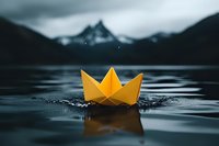 Yellow paper origami boat floating on calm mountain lake water with dramatic cloudy sky and peaks in background creating peaceful minimalist scene.