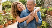 Happy mature Caucasian couple embracing in sunny garden patio with terracotta pots and stone wall. Loving senior man and middle-aged woman sharing joyful moment outdoors.