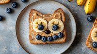 Playful cat-shaped breakfast toast with peanut butter, banana slices as eyes, and blueberries forming smile on gray plate. Creative food art for kids meal.