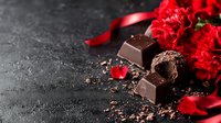Dark chocolate pieces with red carnation flowers and silk ribbon on black textured background, romantic food photography with copy space for Valentine's Day designs.