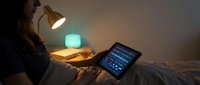 Young woman relaxing in cozy bedroom using tablet device with warm ambient lighting from desk lamp and soft blue glow creating peaceful evening atmosphere.