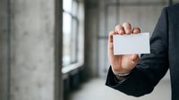 Businessman in dark suit holding blank white business card in modern office corridor with concrete walls and natural window light.