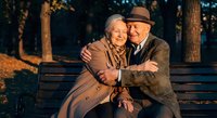 Happy senior couple embracing on park bench during golden hour sunset, showing love and companionship in autumn setting with warm lighting and trees.