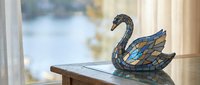 Decorative stained glass swan figurine with blue and amber colors sitting on wooden table near window with soft natural lighting for home decor and crafts.