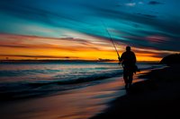 Silhouette of fisherman with fishing rod standing on beach against dramatic sunset sky with vibrant orange and turquoise colors, peaceful ocean waves perfect for travel and outdoor lifestyle designs.