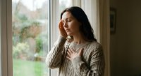 Young woman with long dark hair in cozy sweater touching forehead looking thoughtful by rainy window. Mental health, stress relief, contemplation concept.