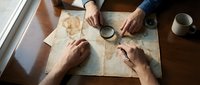 Overhead view of diverse group examining vintage map together at wooden table, planning travel route or adventure with magnifying glass and coffee cup nearby.