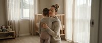 Young Caucasian mother in beige sweater holding toddler son in bright nursery room with white crib. Tender maternal bonding moment for parenting content.