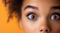 Close up view of brown eyes with long eyelashes and well-groomed eyebrows of young African American woman against vibrant orange background.