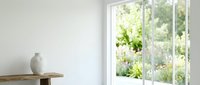 Minimalist interior with white ceramic vase on wooden shelf beside bright window overlooking lush garden, suitable for home decor and lifestyle design projects.