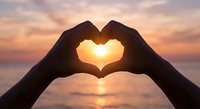 Hands forming heart shape silhouette against golden sunset over ocean water. Romantic gesture symbolizing love, affection and connection during twilight hour.