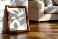 Empty wooden picture frame on hardwood floor with natural sunlight casting leaf shadows through window, white sofa in background, minimalist home decor.