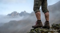 Hiker wearing khaki shorts and hiking boots standing on rocky mountain peak overlooking misty valley landscape during outdoor adventure and trekking expedition.